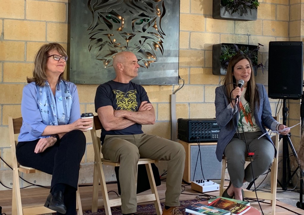 Three people are seated on wooden folding chairs in front of a cinderblock wall and a metal wall hanging carved to resemble the face of a lynx. One person is holding a microphone and speaking. Three books are placed on a small table in front of the three individuals.